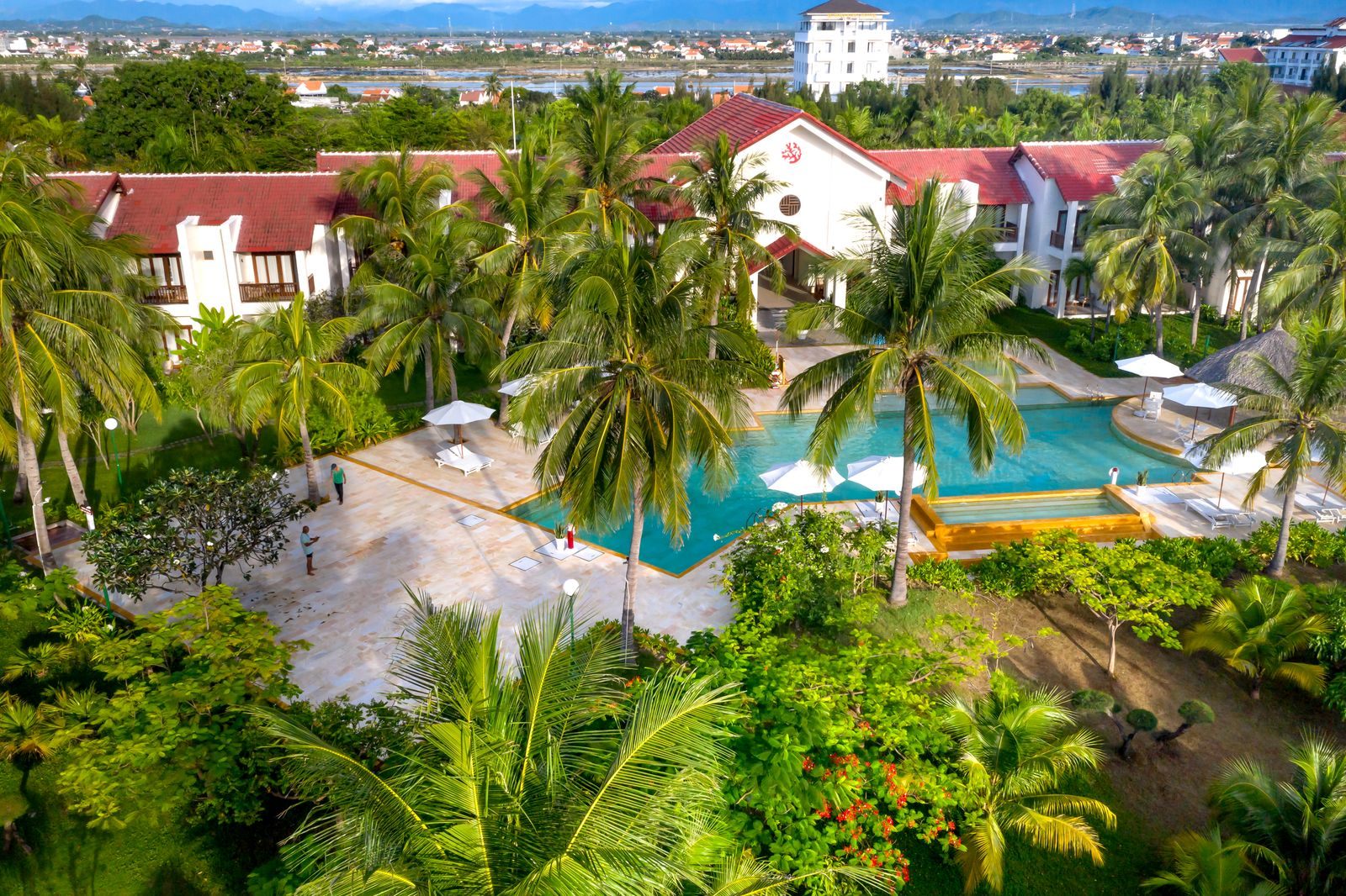 Aerial view of a resort with red-roofed buildings, palm trees and a curving pool, the kind of stay travel clubs advertise